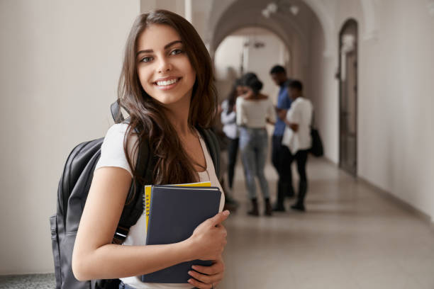Side view of pretty brunette female student posing, standing with notes and backpack on corridor of university. Pretty and clever woman studying in luxury international lyceum.